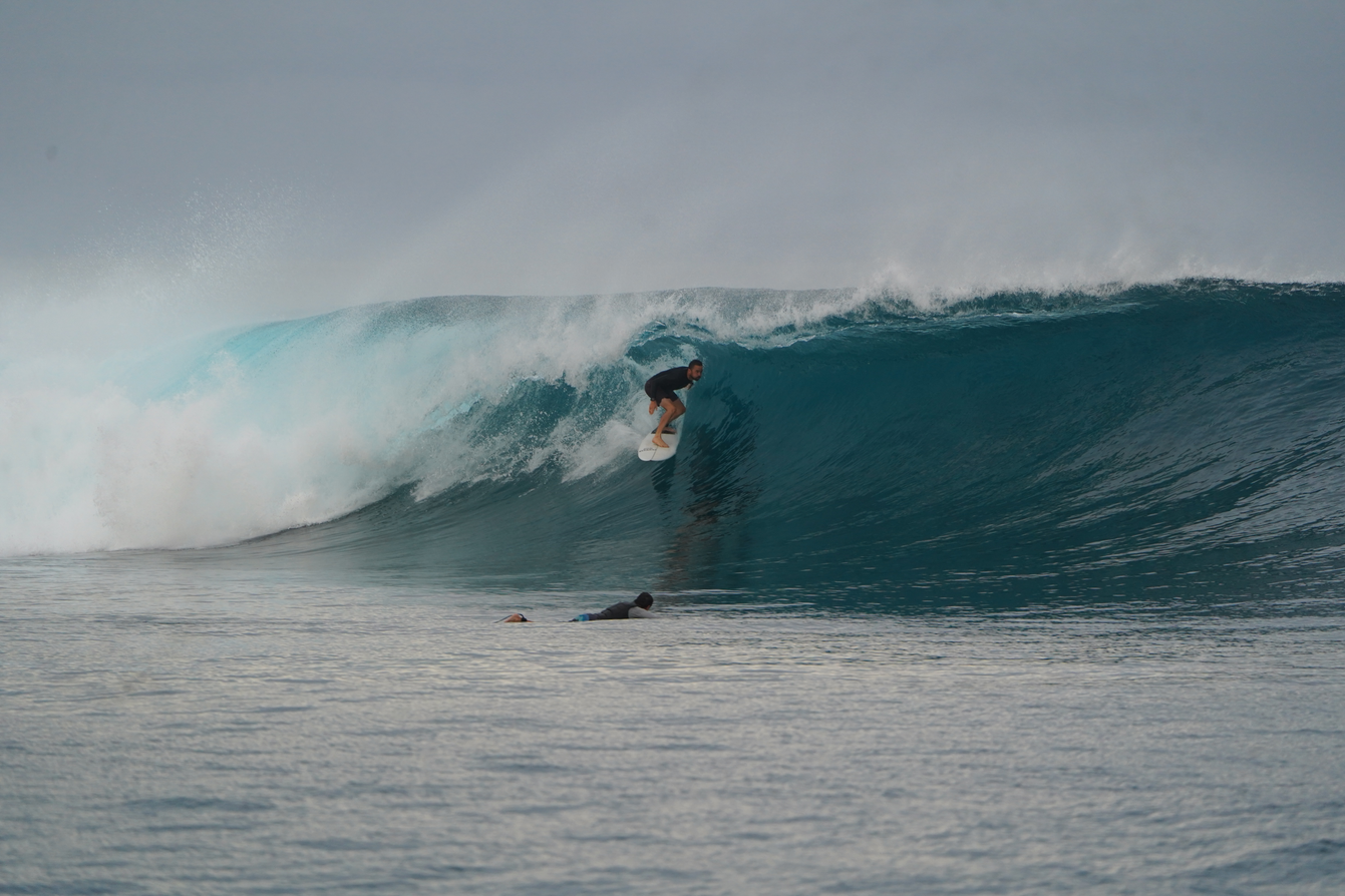 A surfer rides inside a blue wave above another surfer in the water
